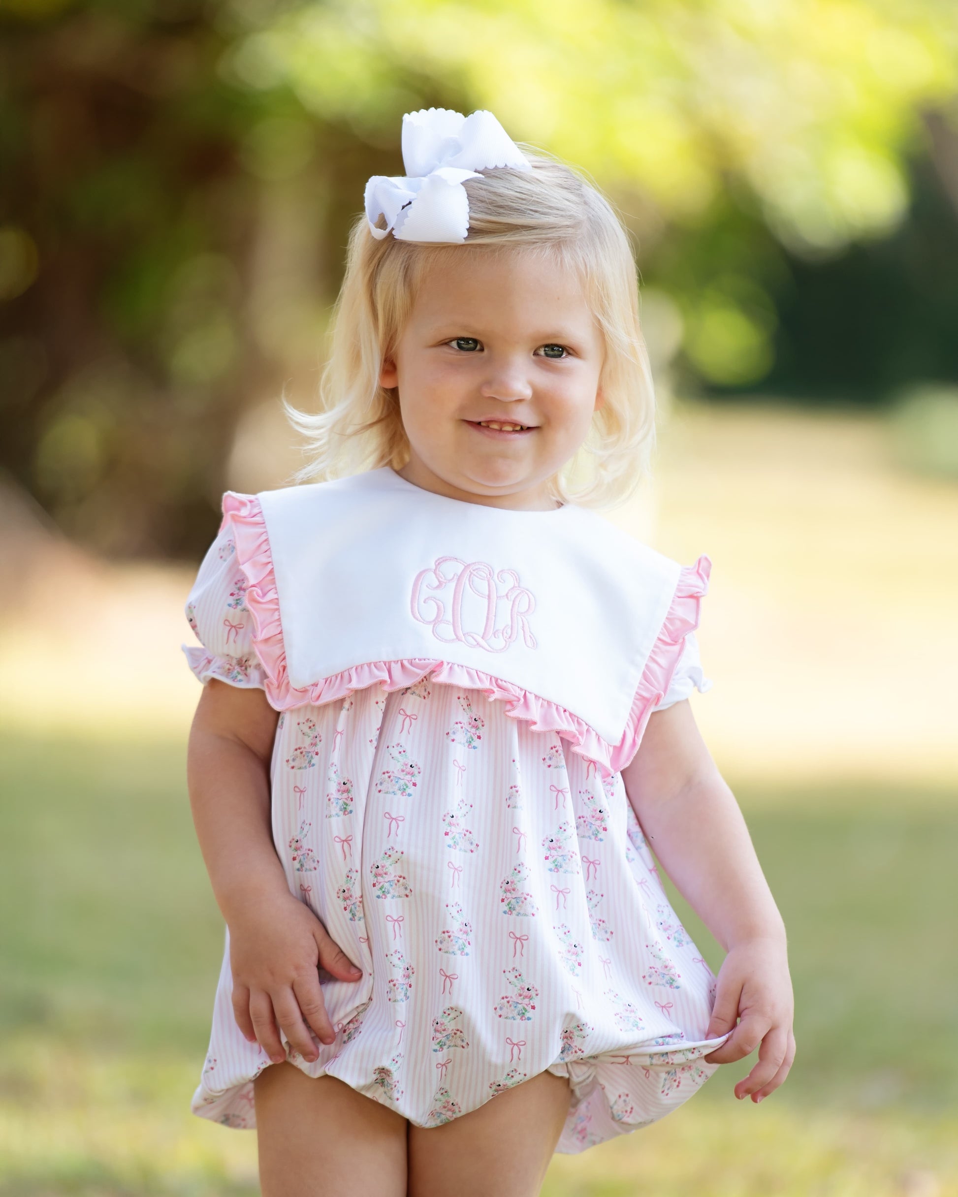Young girl wearing a pink bunny bubble with pink trim and a large bow in her hair, standing outdoors.