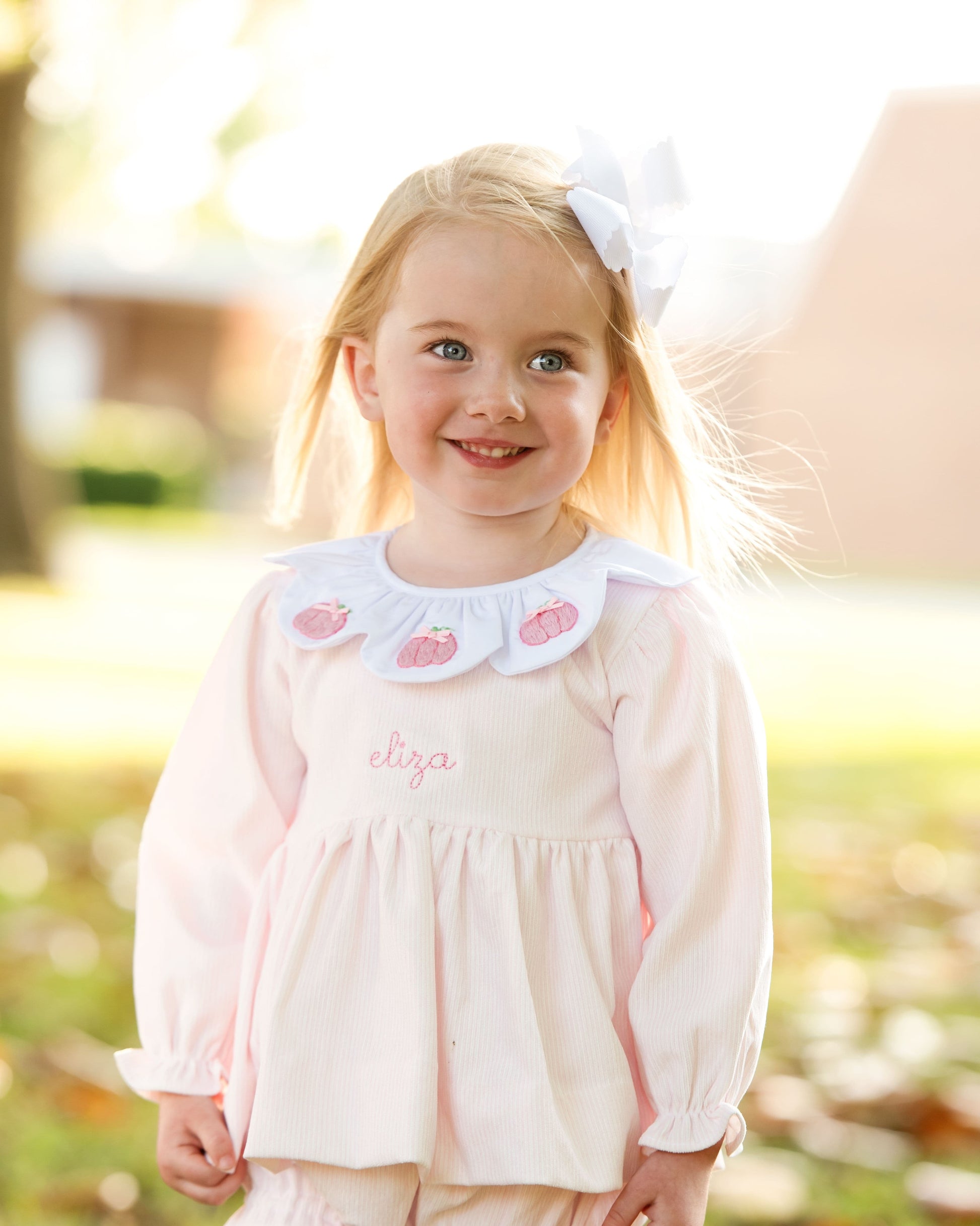 Young girl wearing a pink diaper set with a white collar and embroidered name "eliza" , standing outdoors.