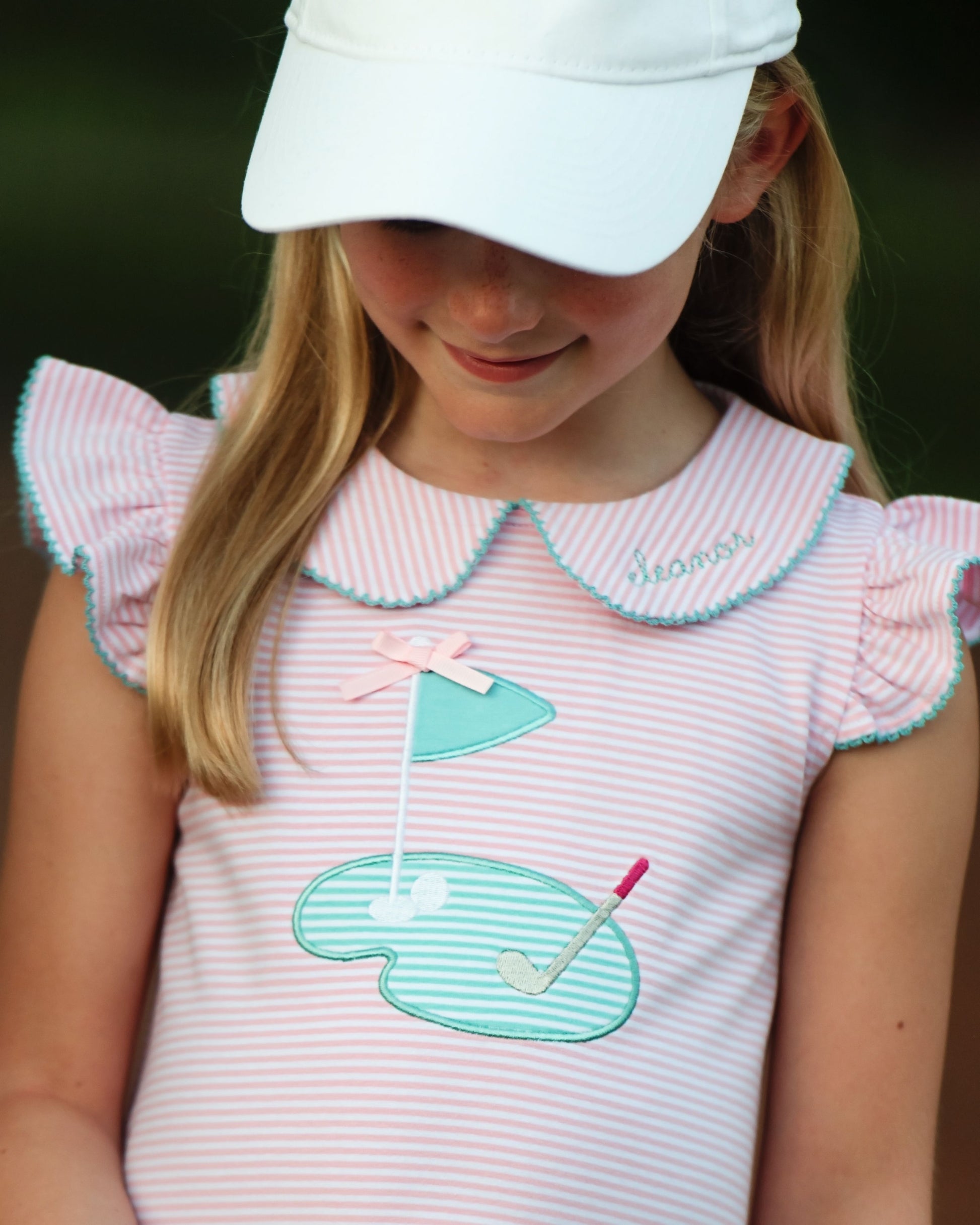 Young girl wearing a pink dress with a golf-themed design and a white cap.