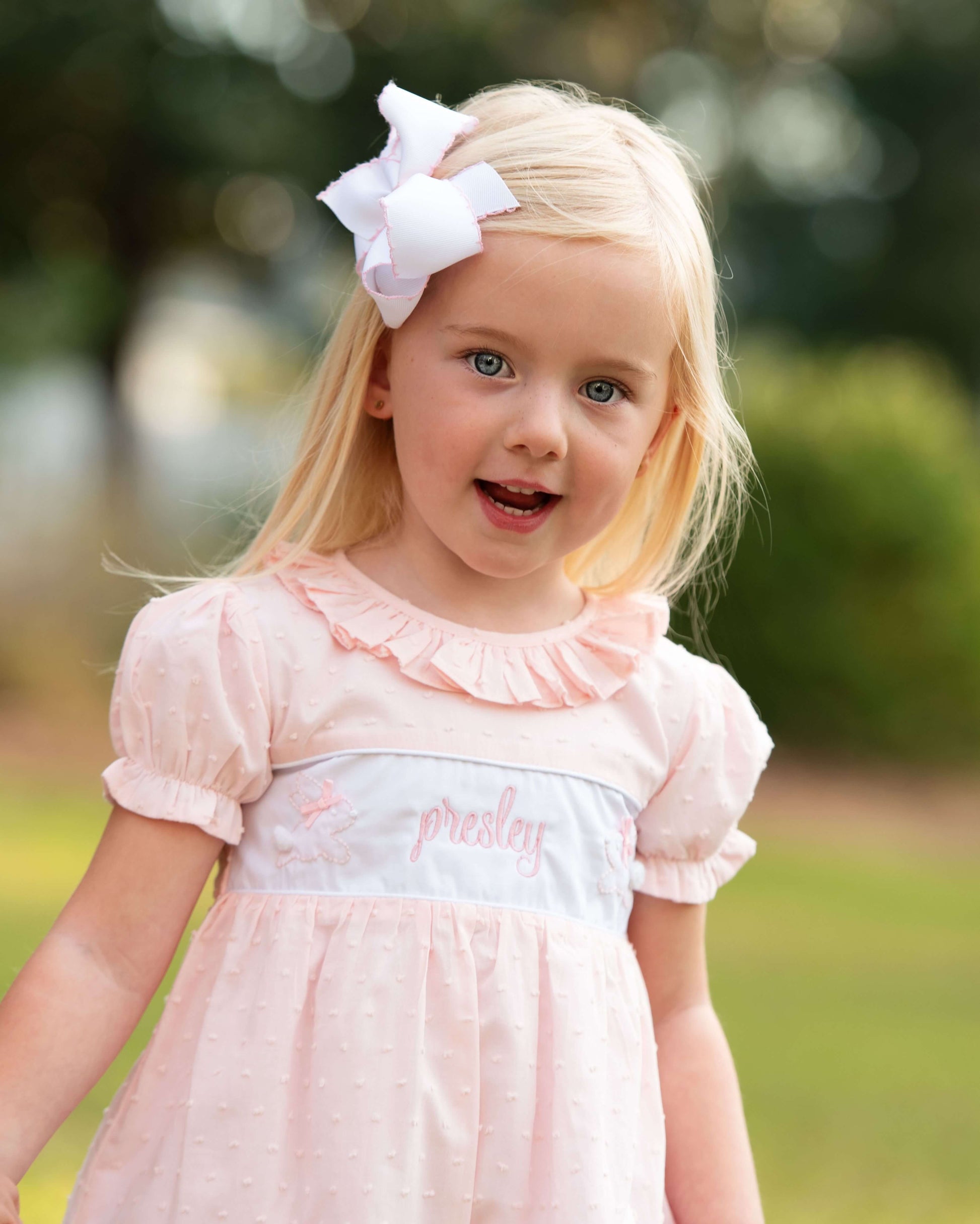 Young girl wearing a pink dress with a white bow in her hair, standing outdoors.