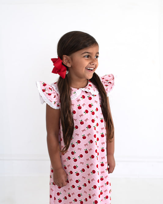 Young girl wearing a pink dress with apples and bows on it. 