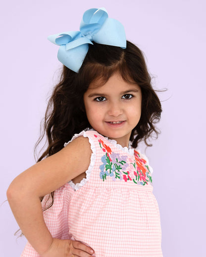 Young girl wearing a pink dress with floral embroidery and a large blue bow in her hair against a light purple background.