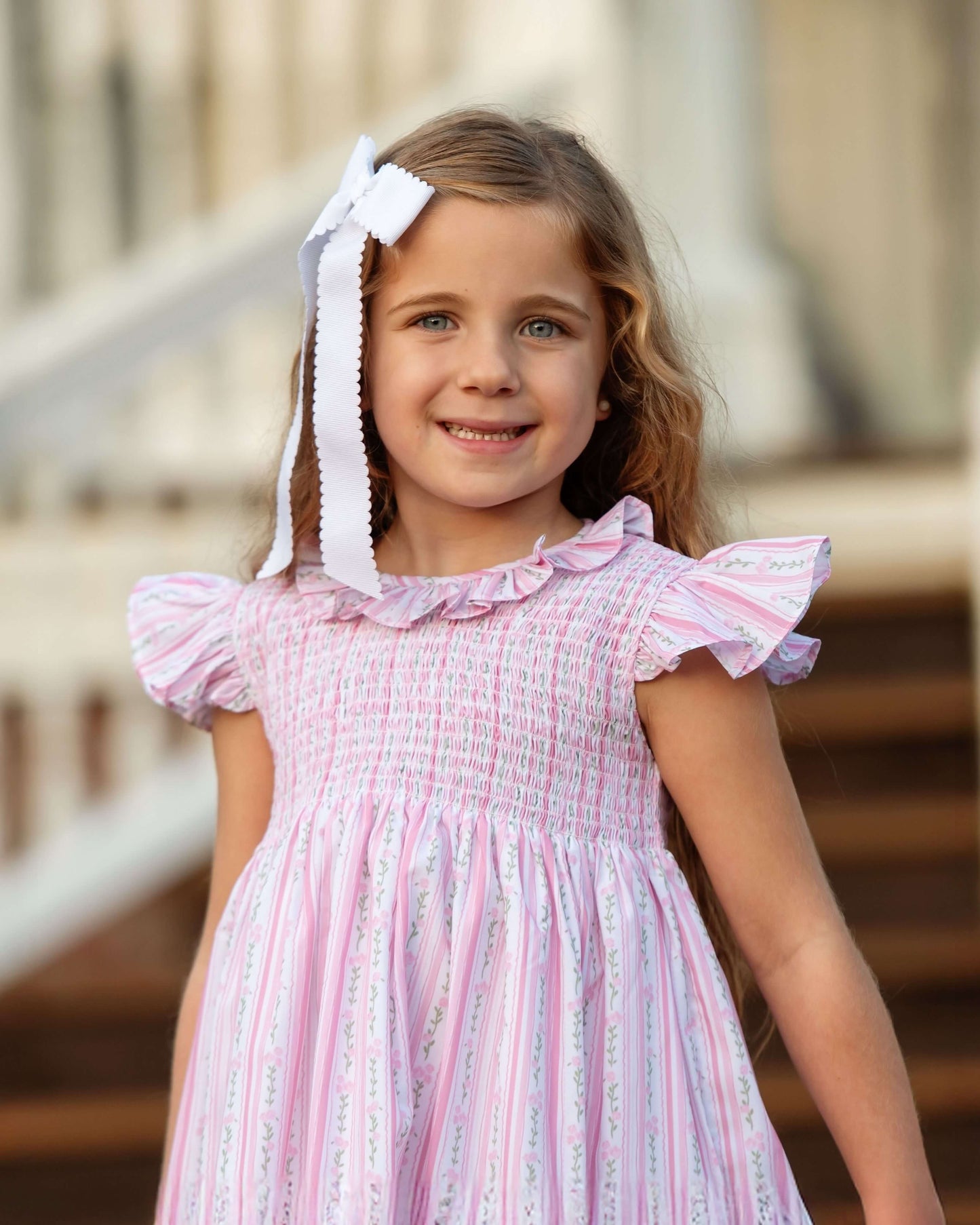 Young girl wearing a pink dress with ruffled sleeves and a white bow in her hair, standing outdoors.