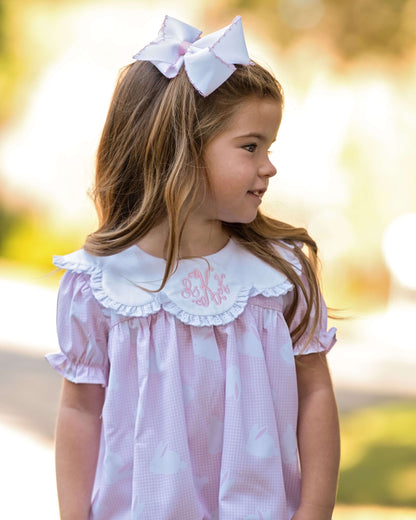 Young girl wearing a pink dress with white collar and embroidered name, outdoors.