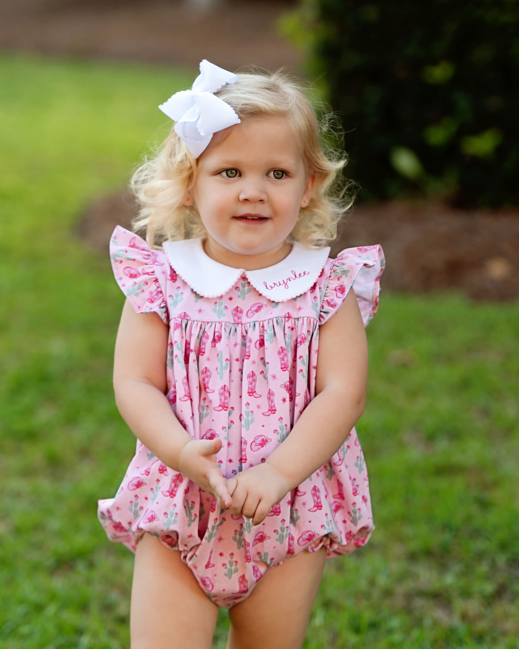 Young girl wearing a pink floral dress with a white collar outdoors on grass.