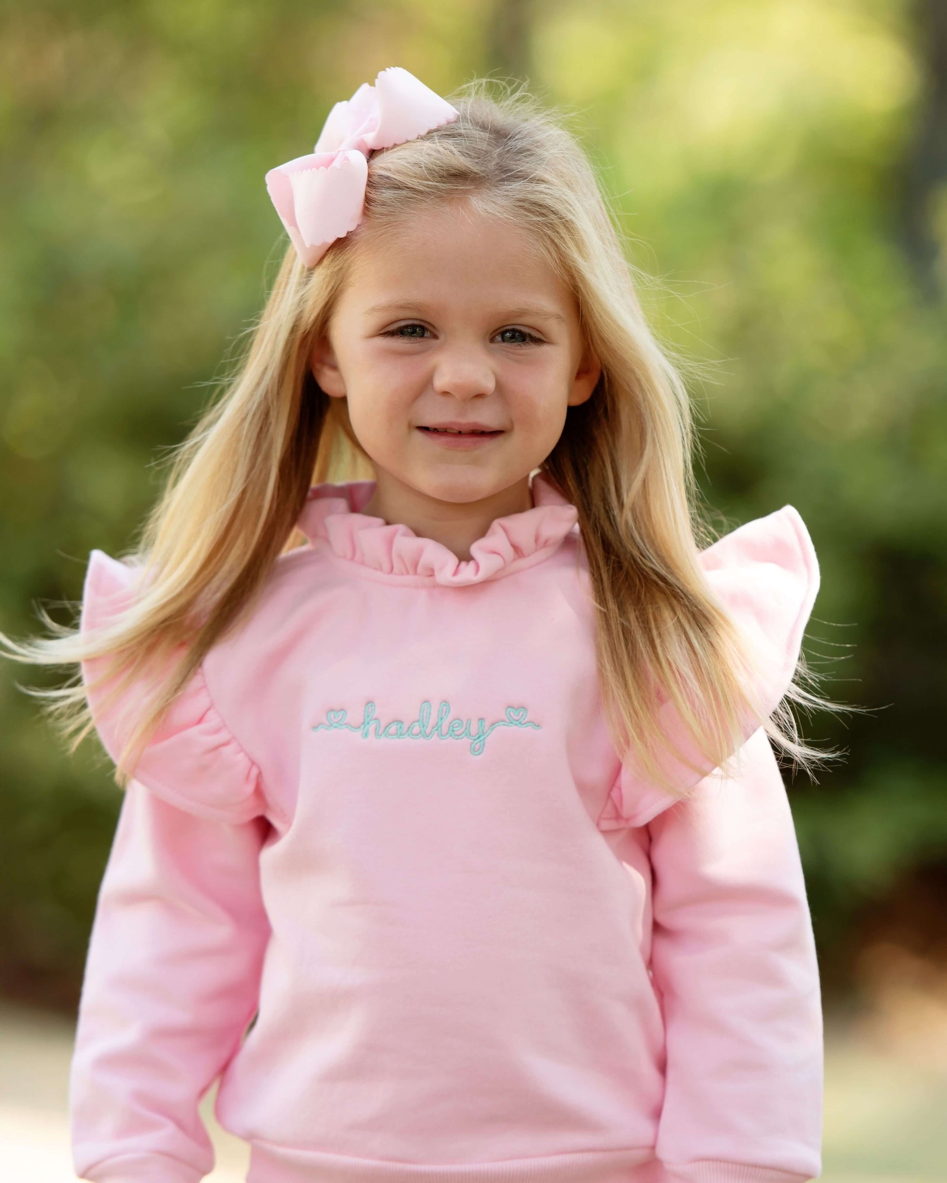 Young girl wearing a pink hoodie with ruffled sleeves and a bow in her hair, standing outdoors.