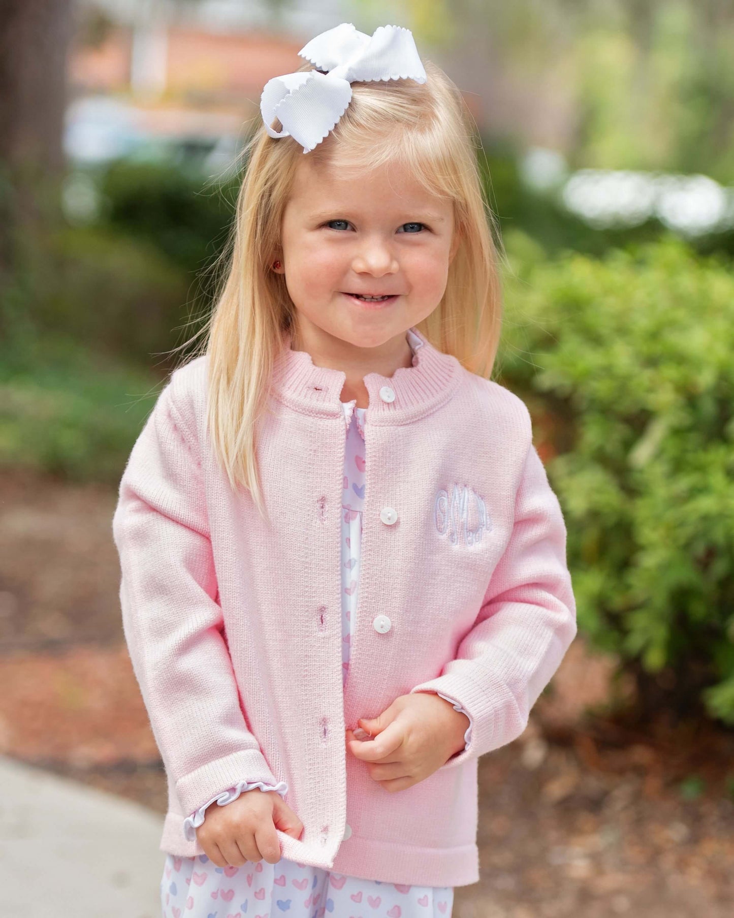 Young girl wearing a pink jacket with a white bow in her hair, standing outdoors.