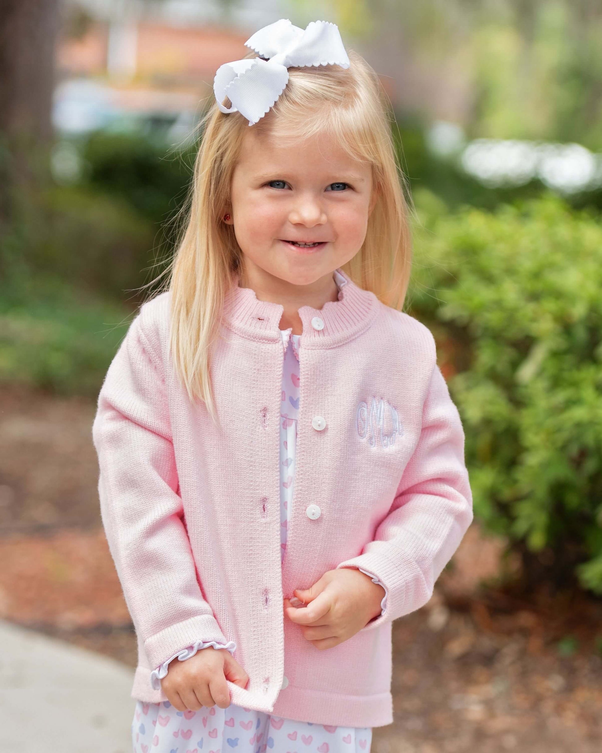 Young girl wearing a pink jacket with a white bow in her hair, standing outdoors.