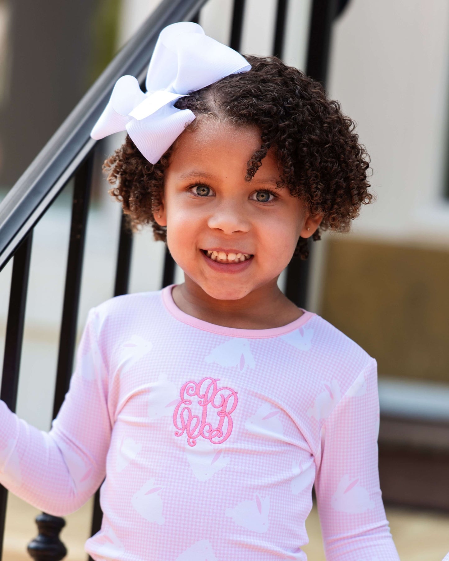 Young girl wearing a pink long-sleeve shirt with a bow in her hair, standing outdoors.