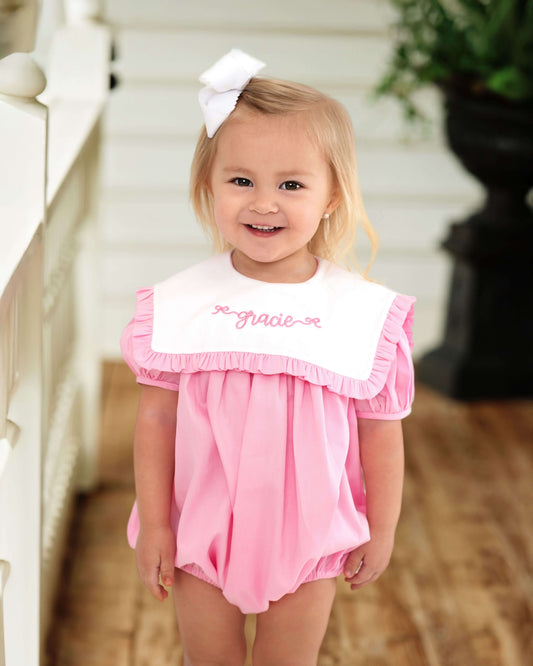 Young girl wearing a pink romper with white ruffled collar and 'First Grade' text, standing outdoors.