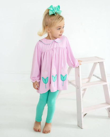 Young girl wearing a pink set with green tulip designs and green leggings, standing next to a white stool.