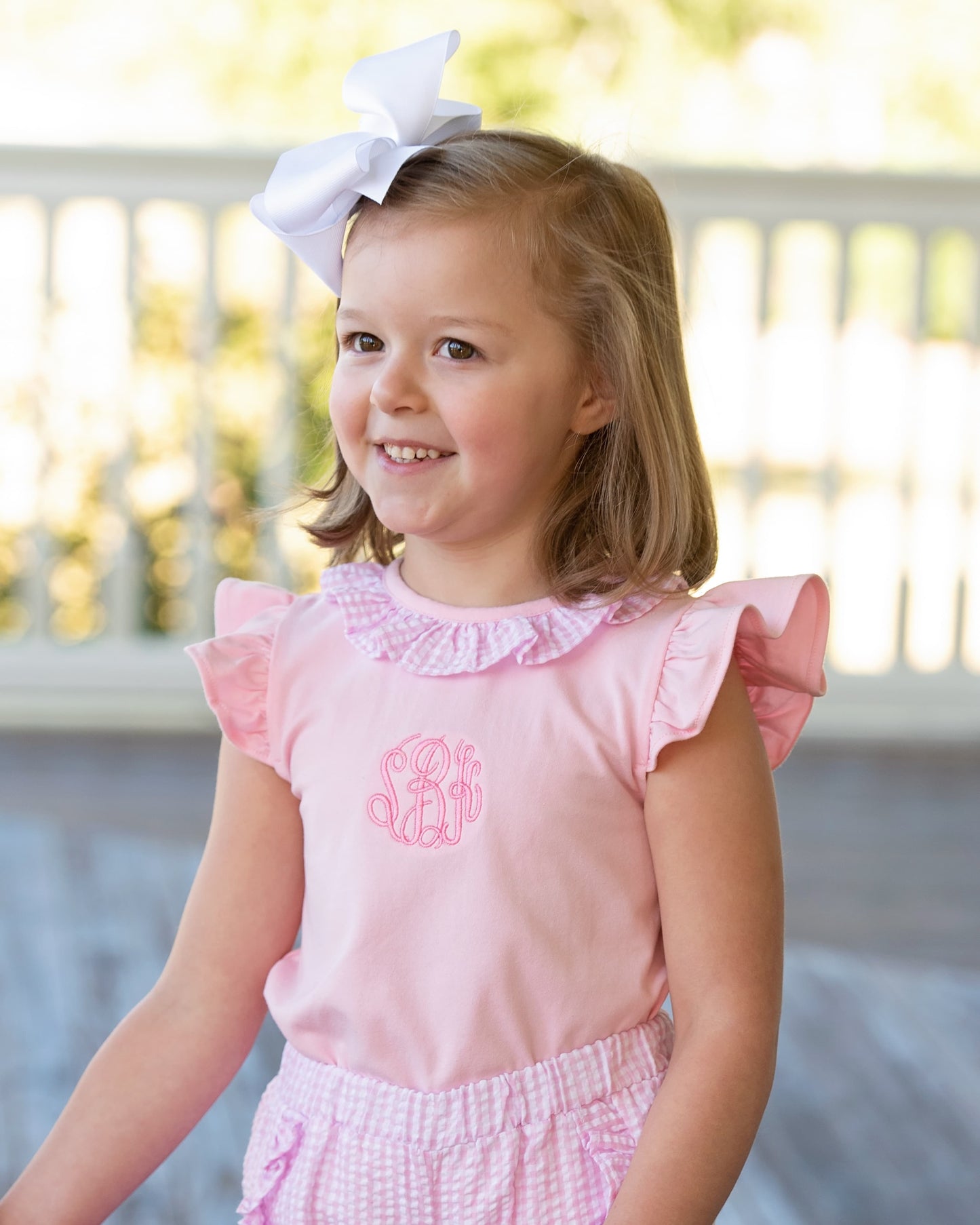 Young girl wearing a pink short set with ruffled sleeves and a white bow in her hair, standing on a wooden deck.