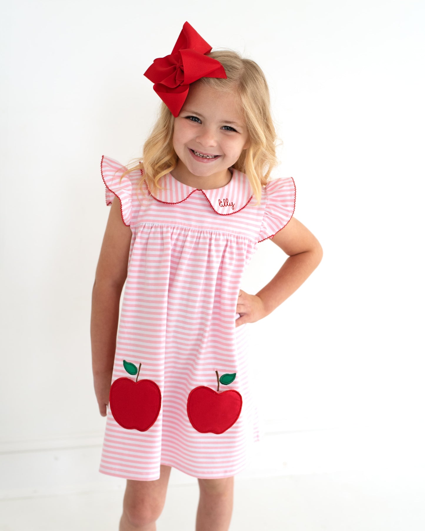 Young girl wearing a pink striped dress with apple pockets and a red bow on her hair.