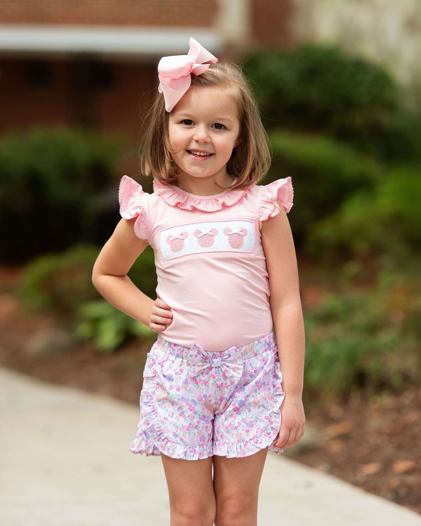 Young girl wearing a pink top and floral shorts with a large bow in her hair, standing outdoors.