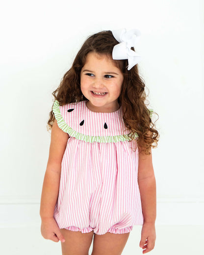 Young girl wearing a pink watermelon-themed romper with a white bow in her hair on a white background