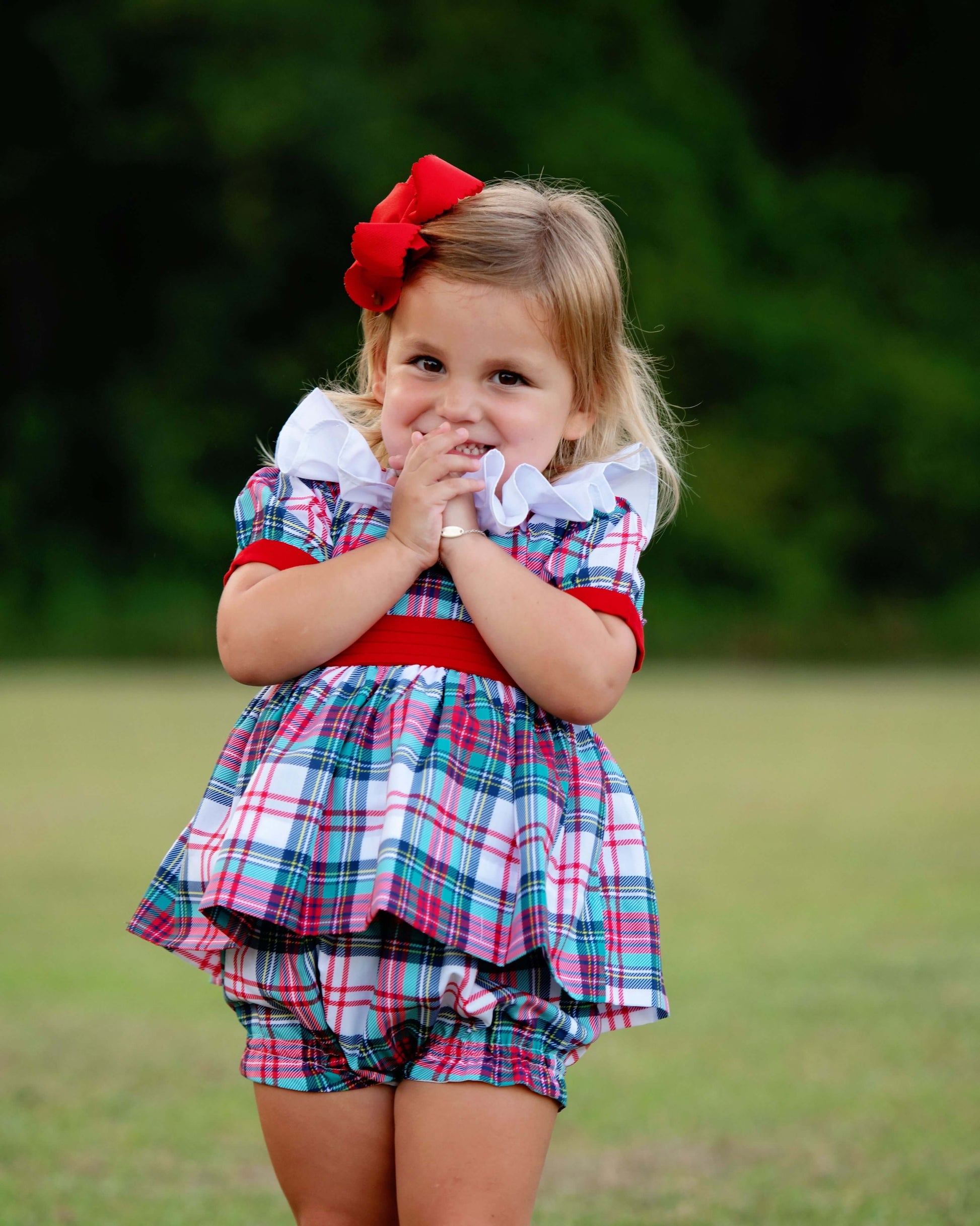 Young girl wearing a plaid diaper set with a smocked ruffled collar and a red bow in her hair, standing outdoors.