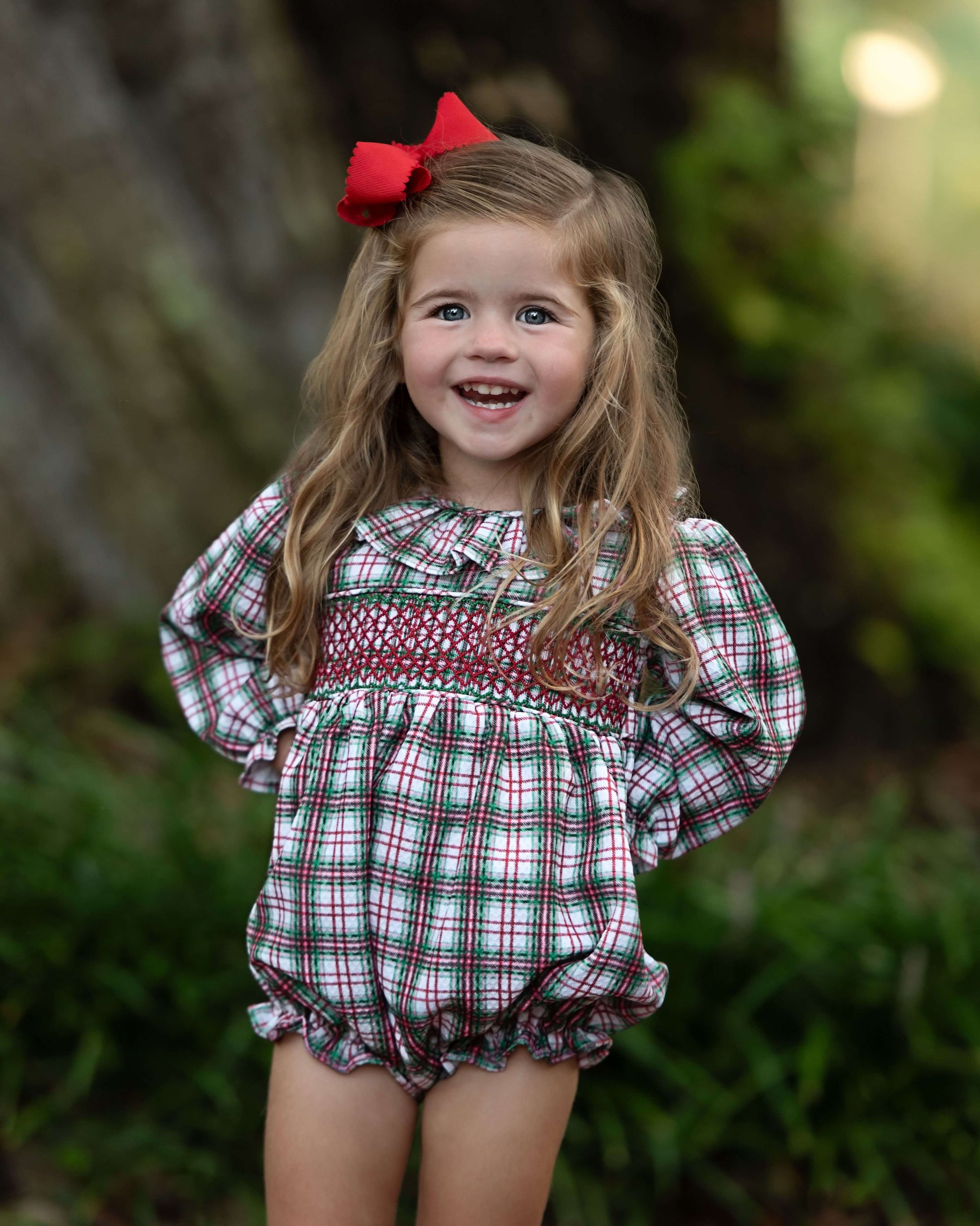 Young girl wearing a plaid dress with a red bow in her hair, standing outdoors.