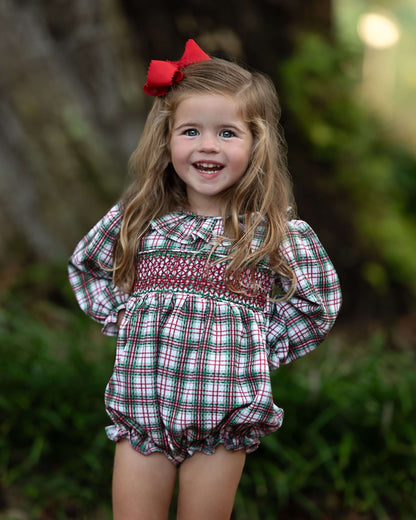 Young girl wearing a plaid dress with a red bow in her hair, standing outdoors.