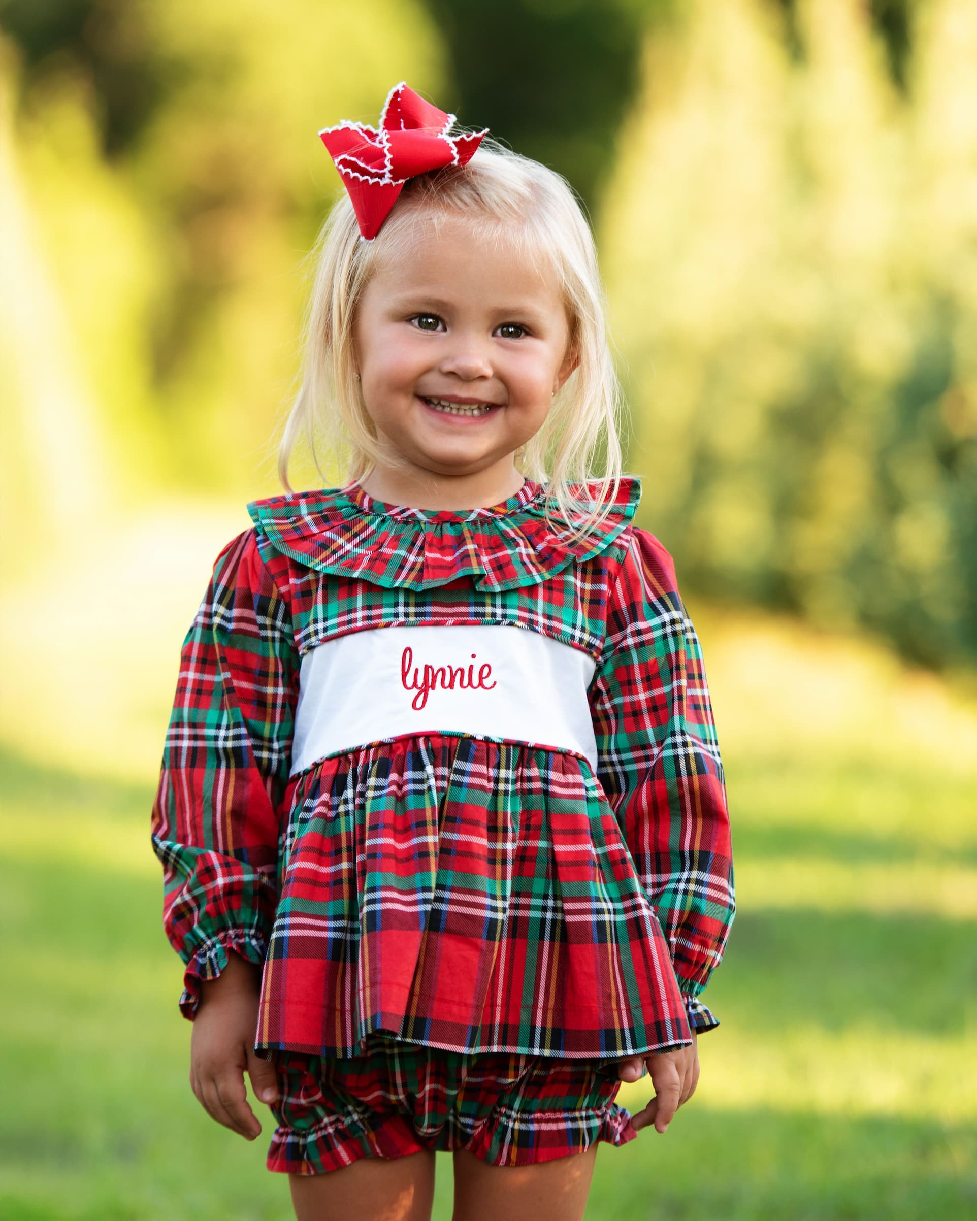 Young girl wearing a red and green plaid dress with a name tag, standing outdoors.
