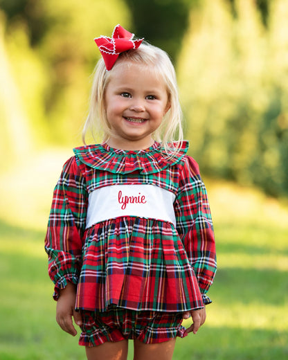 Young girl wearing a red and green plaid dress with a name tag, standing outdoors.