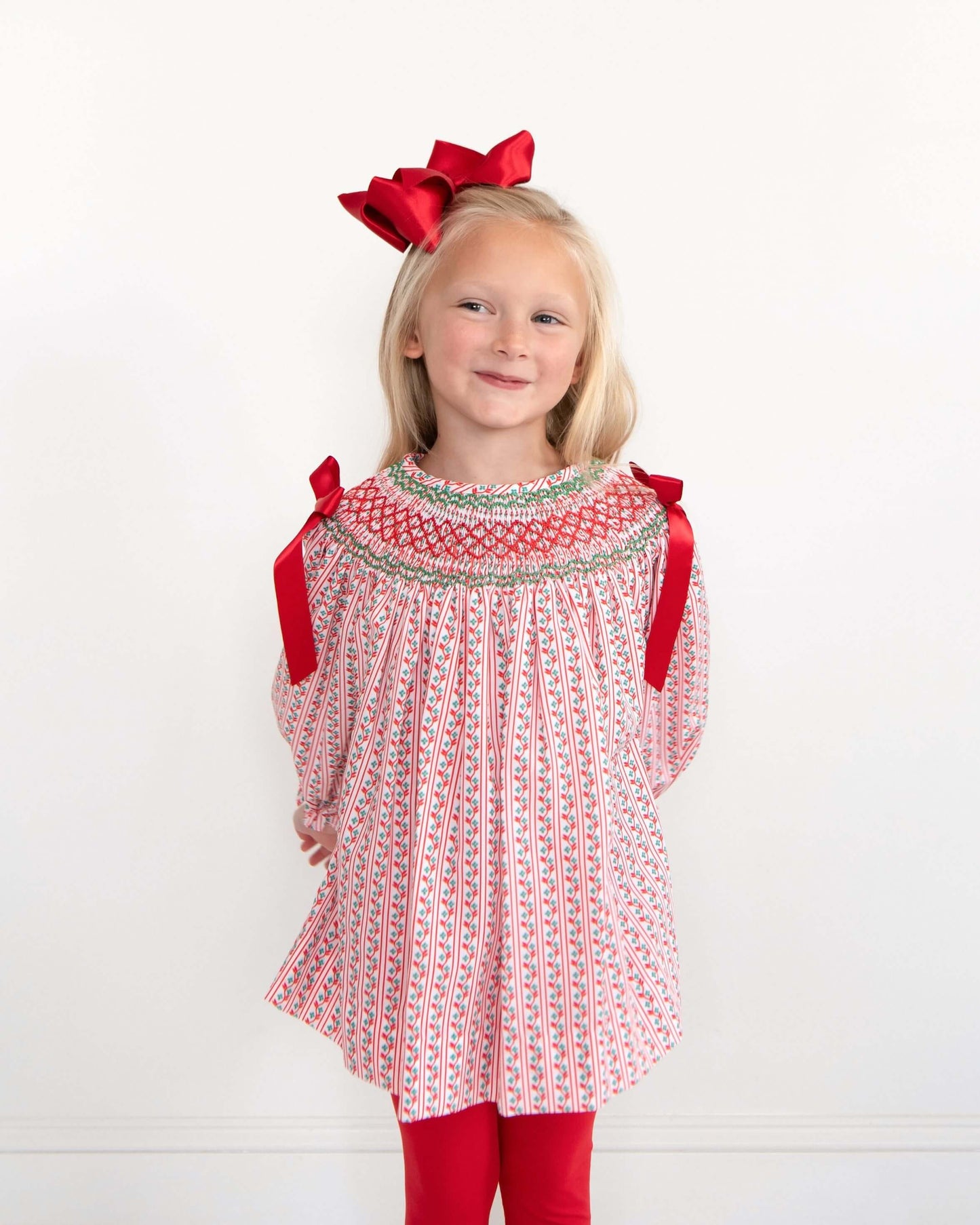 Young girl wearing a red and white patterned long sleeved top with red bows and matching red leggings on a white background