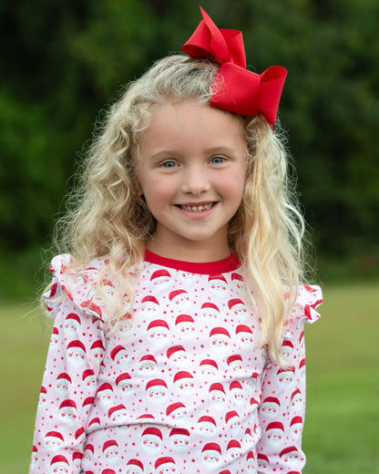 Young girl wearing a red and white patterned outfit with a large red bow in her hair, standing outdoors.