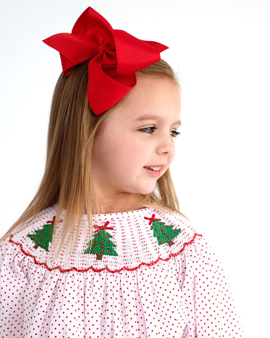 Young girl wearing a red bow and a red dot dress with smocked Christmas tree designs on a white background