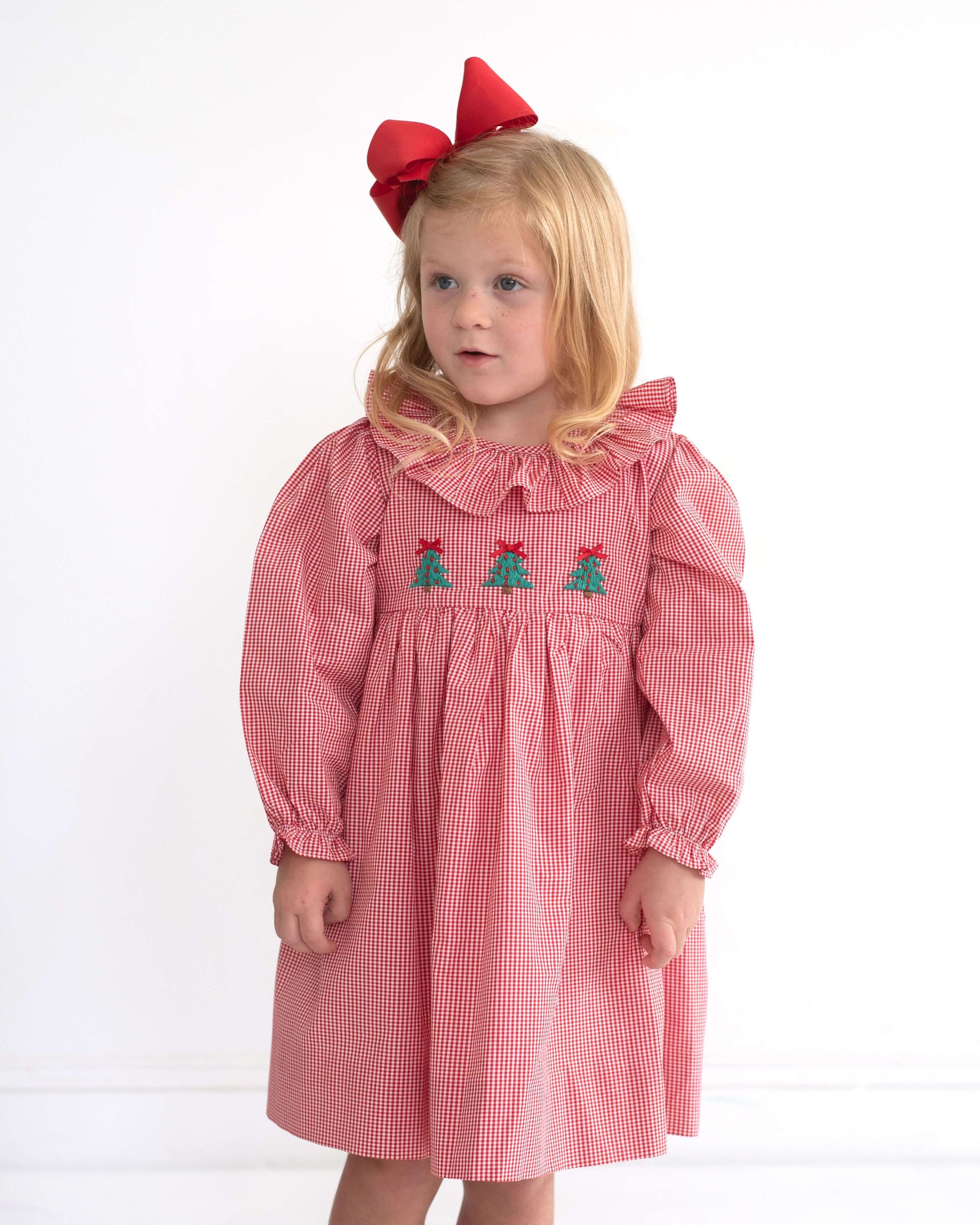 Young girl wearing a red checkered dress with green tree designs and a large red bow on a white background