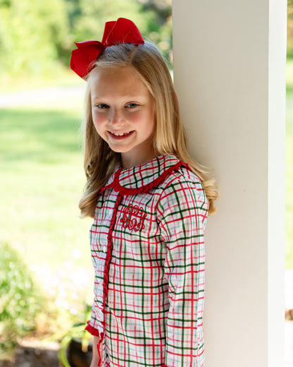 Young girl wearing a red, green, and white plaid shirt with a large red bow in her hair, standing outdoors.