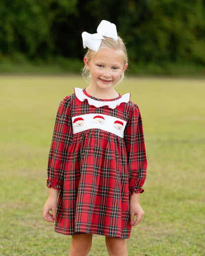 Young girl wearing a red plaid dress with white collar and Santa face design, standing outdoors.