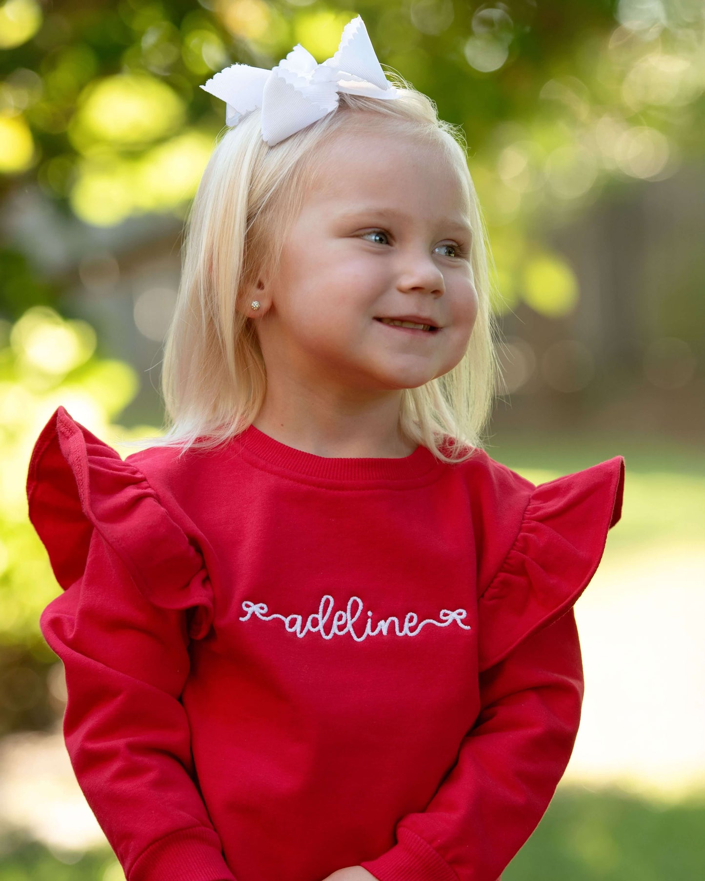 Young girl wearing a red sweatshirt with 'Madeline' on it, outdoors.
