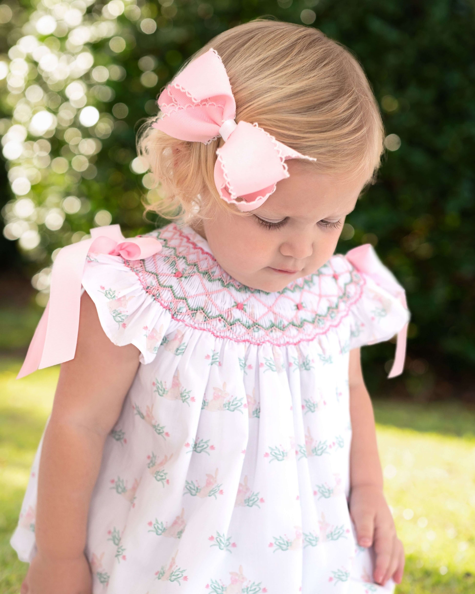 Young girl wearing a smocked floral bubble with pink ribbons and a large bow outdoors.