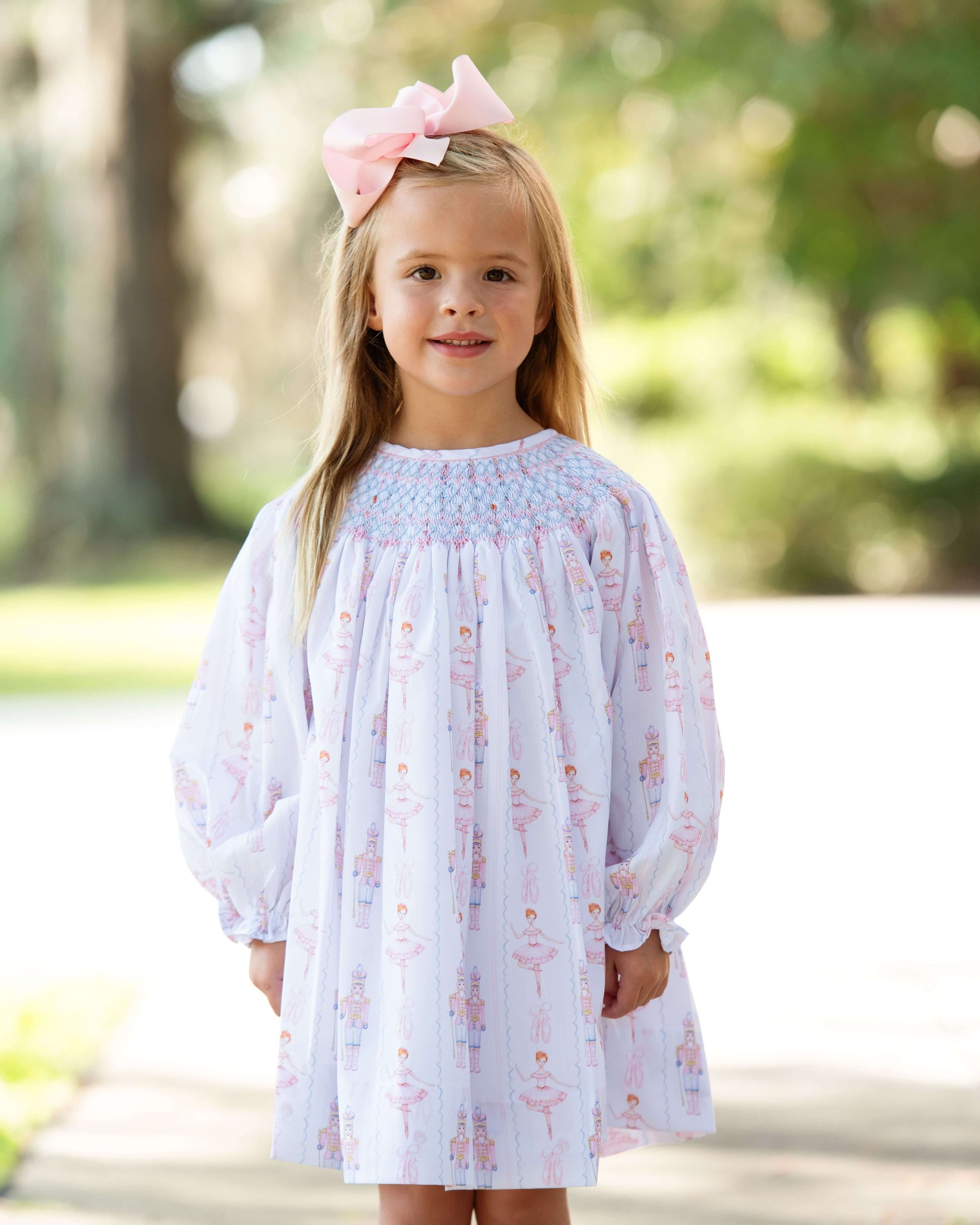 Young girl wearing a smocked holiday print dress with a pink bow in her hair, standing outdoors.