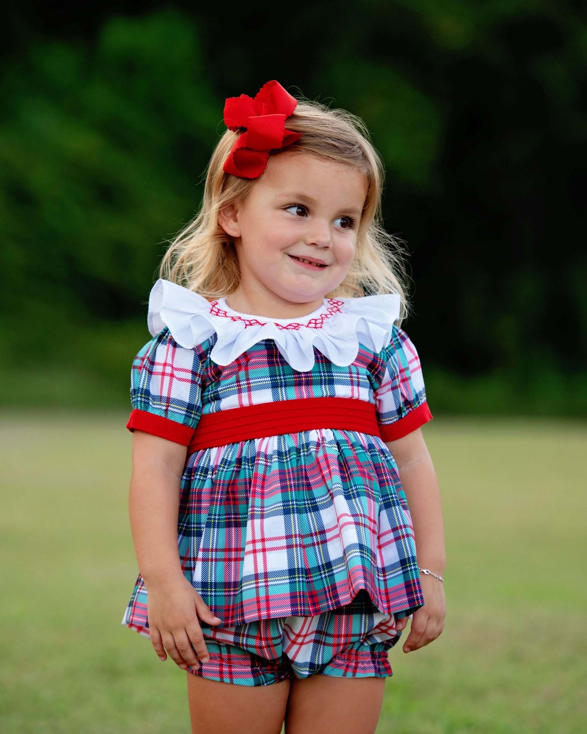 Young girl wearing a smocked plaid diaper set with a red ribbon in her hair, standing outdoors.