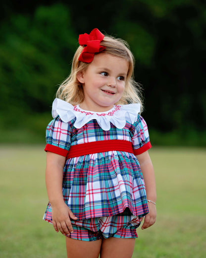 Young girl wearing a smocked plaid diaper set with a red ribbon in her hair, standing outdoors.