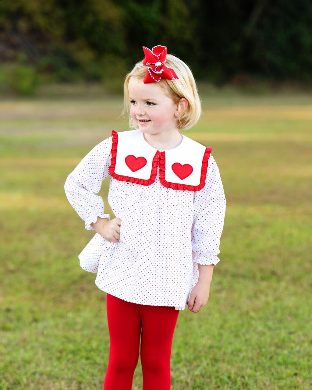 Young girl wearing a white blouse with red heart details and red leggings standing in a grassy field.