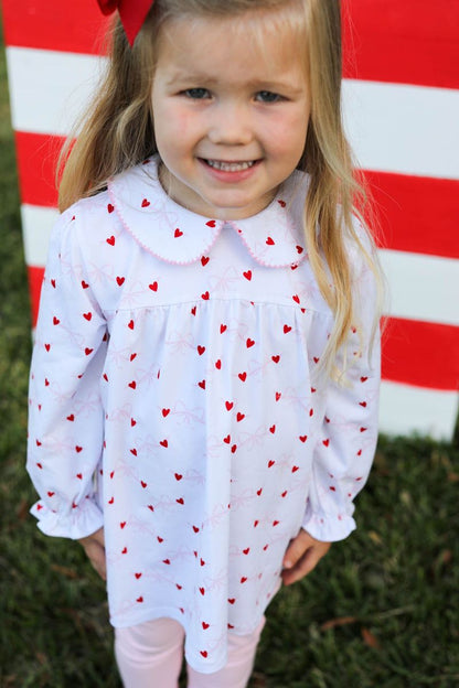 Young girl wearing a white blouse with red heart patterns in front of a red and white striped background.