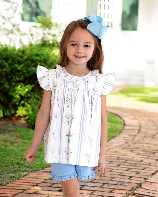 Young girl wearing a white blouse with ruffled sleeves and blue shorts, standing on a brick path with greenery in the background.