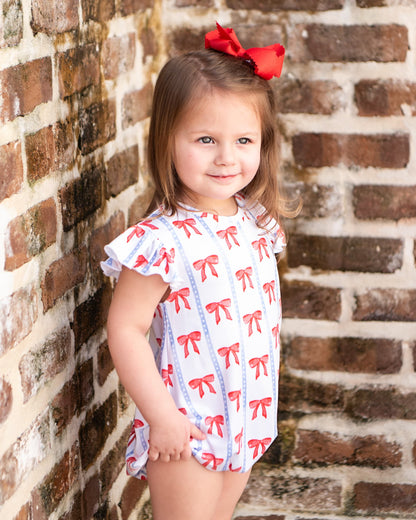 Young girl wearing a white bubble with red bows against a brick wall.