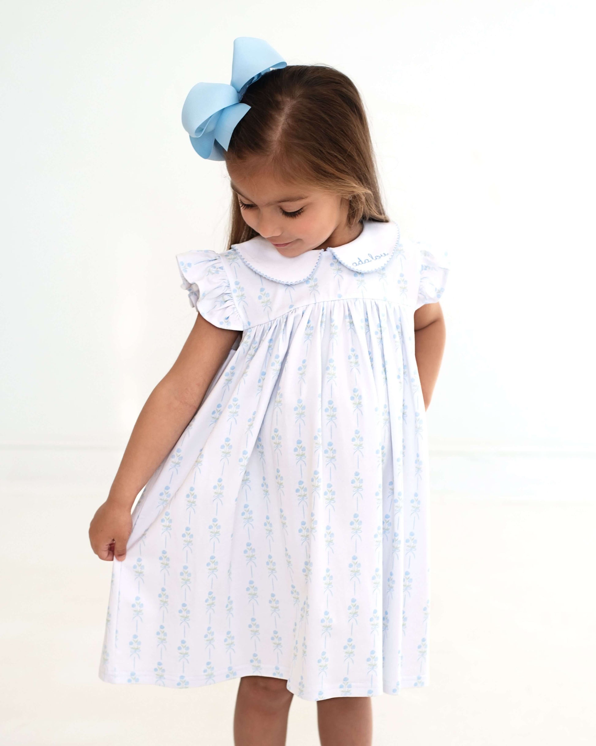 Young girl wearing a white dress with blue floral patterns and a large blue bow in her hair on a white background.