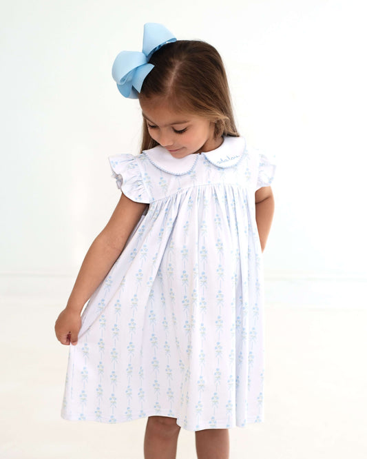 Young girl wearing a white dress with blue floral patterns and a large blue bow in her hair on a white background.
