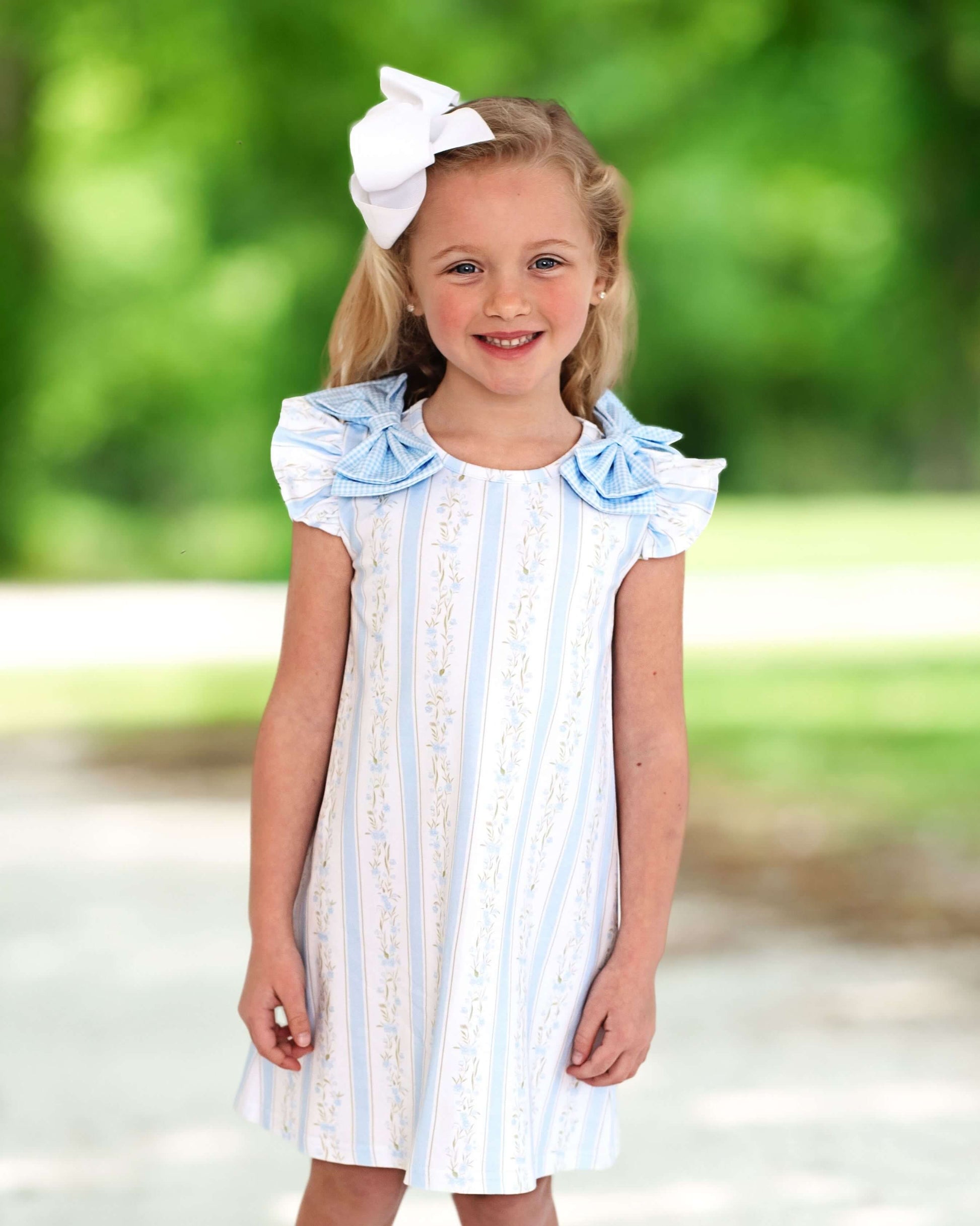 Young girl wearing a white dress with blue stripes and ruffled sleeves, standing outdoors with a blurred green background.