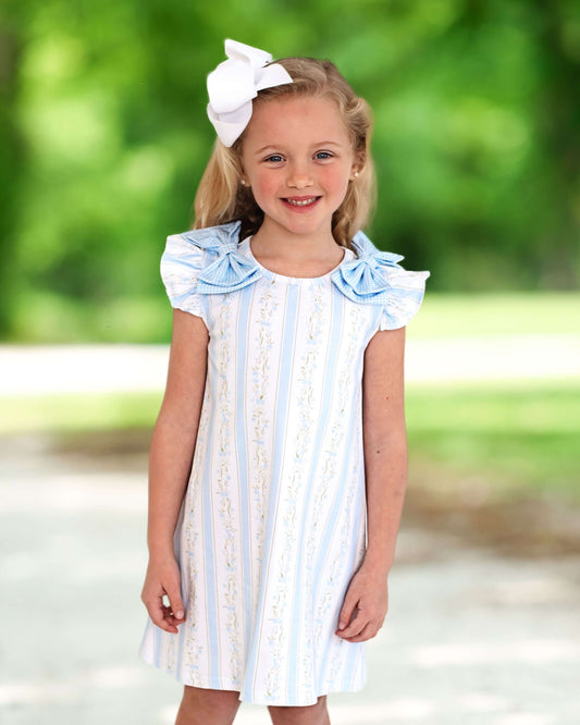 Young girl wearing a white dress with blue stripes and ruffled sleeves, standing outdoors with a blurred green background.