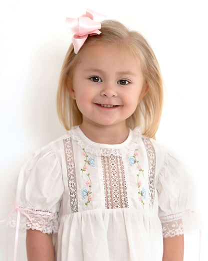Young girl wearing a white dress with floral embroidery and a pink bow in her hair on a white background