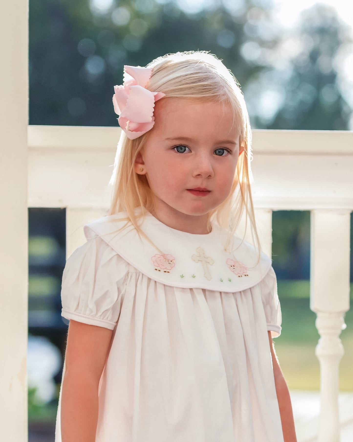 Young girl wearing a white dress with floral embroidery and a pink bow in her hair, standing outdoors.