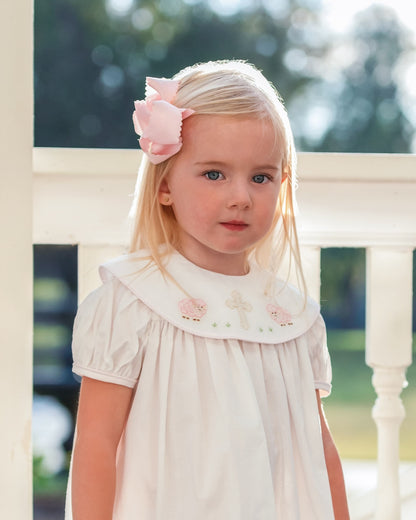 Young girl wearing a white dress with floral embroidery and a pink bow in her hair, standing outdoors.