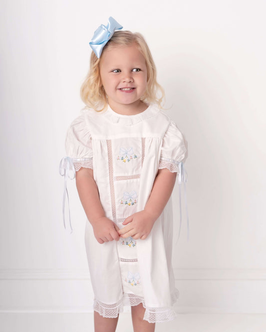 Young girl wearing a white dress with lace details and a blue bow in her hair on a white background