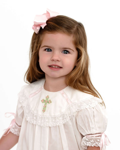 Young girl wearing a white dress with lace details and a pink bow in her hair on a white background