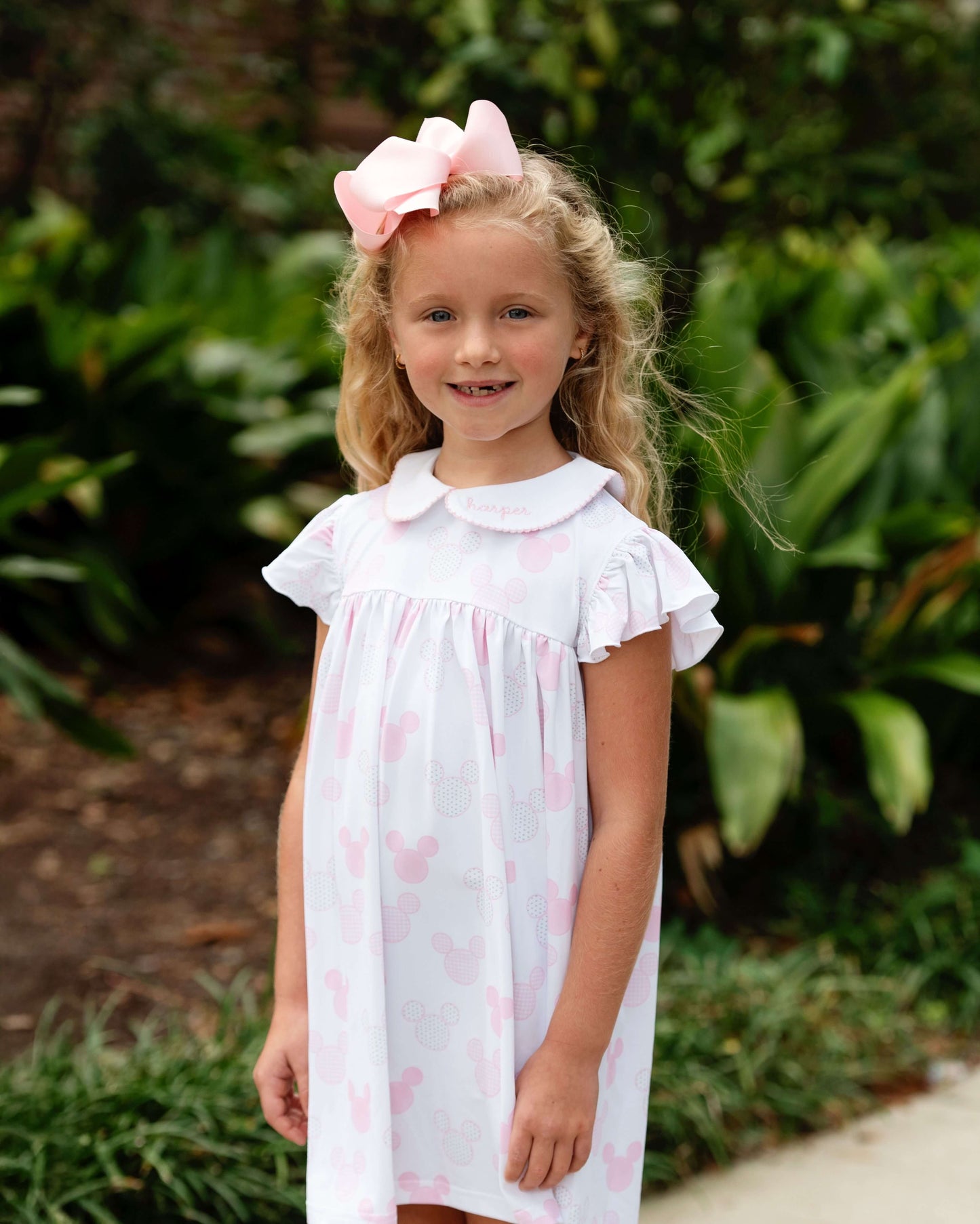 Young girl wearing a white dress with pink hearts and a large pink bow outdoors.