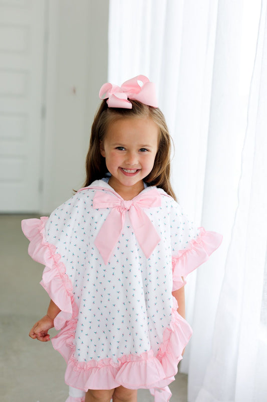 Young girl wearing a white dress with pink ruffles and a bow, standing in front of white curtains.
